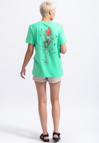 Muntgroene katoenen T-shirt met korte mouwen, met een speelse grafiek op de achterkant van een figuur met een rode appel als hoofd en ranken.