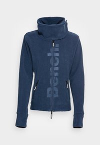 Navy blue fleece jacket with high collar, zip closure, and two zipped pockets. Features a large tonal logo down the front.