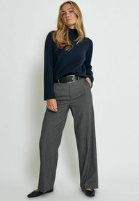 Maglione blu navy a maniche lunghe, abbinato a pantaloni larghi grigi chiari. Cintura nera con fibbia rettangolare che accentua la vita. Ballerine nere.