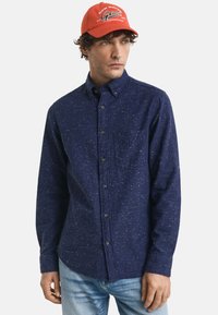 Marineblauw overhemd met knopen en een gespikkeld patroon, borstzak en lange mouwen. Gecombineerd met een feloranje pet met logo-tekst.