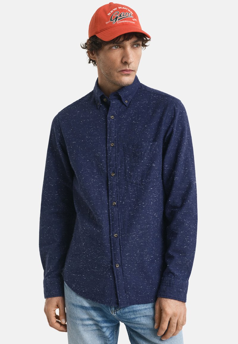 Marineblauw overhemd met knopen en een gespikkeld patroon, borstzak en lange mouwen. Gecombineerd met een feloranje pet met logo-tekst.