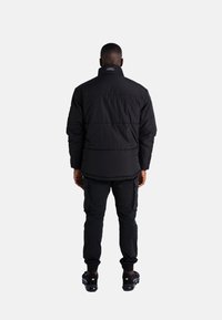 Mann in schwarzer gepolsterter Jacke, schwarzen Cargo-Hosen und schwarzen Sneakers, der mit dem Rücken zu einem schlichten weißen Hintergrund steht.