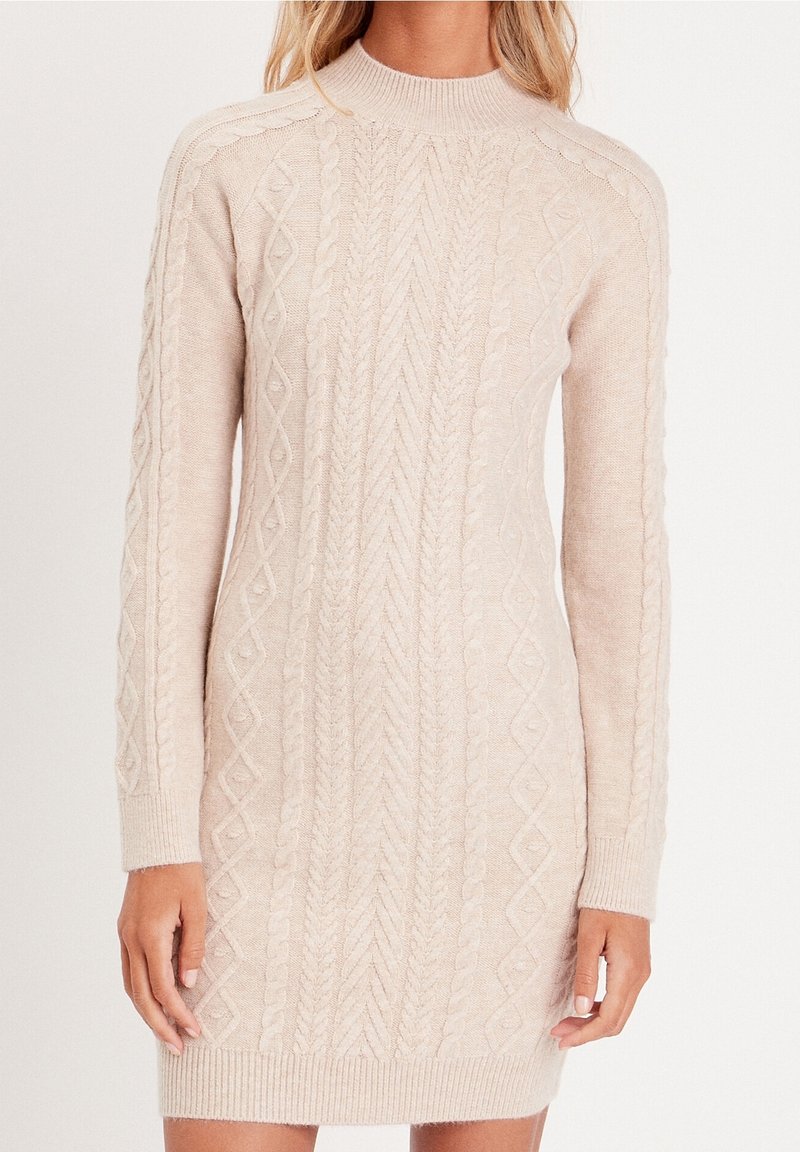 Femme portant une robe-pull beige en maille torsadée avec manches longues et poignets côtelés, debout devant un fond uni.