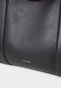Borsa a mano in pelle nera con finitura testurizzata, con logo dorato "Calvin Klein" e linee pulite, che enfatizzano un design elegante.