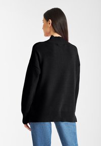 Schwarzer oversized Pullover mit einem gerippten hohen Kragen und Bündchen, aus einem glatten Material mit lässigem Schnitt, kombiniert mit hellblauen Jeans.