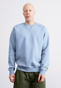 Lys blå sweatshirt med ribbede manchetter og kant, med rund halsudskæring og en subtil logodetalje på ærmet. Bæres over olivengrønne bukser.
