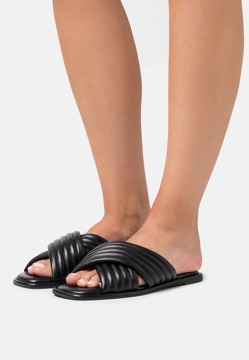 Pieds portant des sandales plates noires matelassées avec des lanières croisées, bout ouvert et design à enfiler sur un fond blanc.