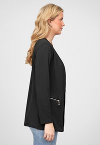 Svart teksturert jakke med lange ermer og glidelås-lomme på siden, med rett snitt og avslappet passform, båret over blå jeans.