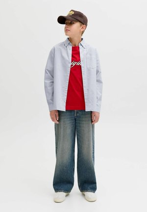 Fiú áll, barna sapkát, piros grafikus pólót, világoskék, gombos ing nyitva, bőszárú, koptatott farmert és fehér sportcipőt visel.