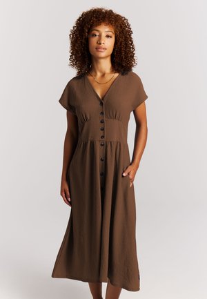 Vrouw met krullend haar die een bruine midi-jurk met knopen, korte mouwen en zakken draagt, staand tegen een effen lichte achtergrond.