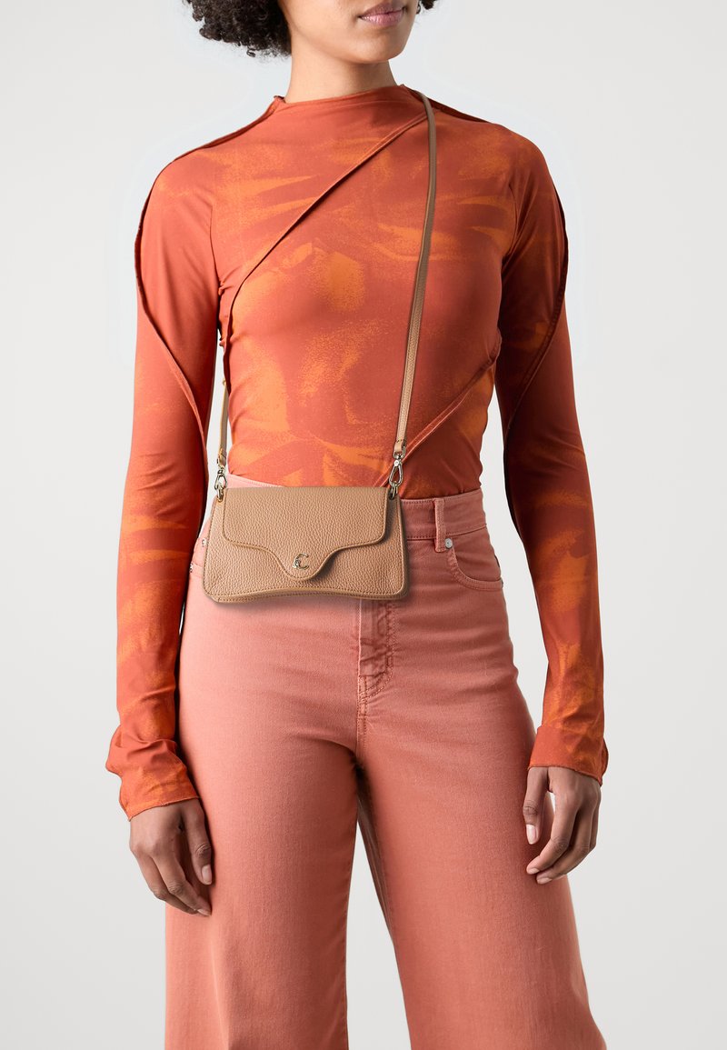 Hnědá crossbody kabelka s texturovaným povrchem, zakřivenou klopou a kovovými detaily, nošená přes dlouhý top s oranžovým vzorem a růžové džíny.