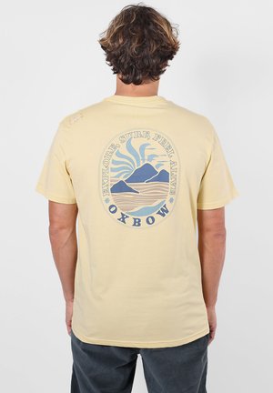 Personne aux cheveux bruns courts portant un t-shirt jaune pâle avec un graphisme bleu de montagne et de soleil ainsi qu’un texte au dos.