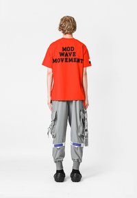 Camisa roja de manga corta con el texto negro "MOD WAVE MOVEMENT" en la parte posterior; combinada con pantalones cargo grises holgados que presentan múltiples bolsillos y correas.