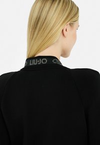 Vêtement noir avec une texture lisse, doté d'un col à motif dans une couleur contrastante. Le col comprend des détails textuels réfléchissants.