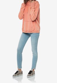 Felpa pesca con tasca a marsupio, indossata con jeans skinny blu chiaro e sneaker con stampa serpente caratterizzate da accenti neri e suola piatta.