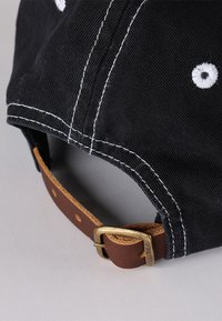 Fekete pamutsapka fehér varrással és hímzett szellőzőlyukakkal. Állítható barna bőrpánt réz csattal hátul.