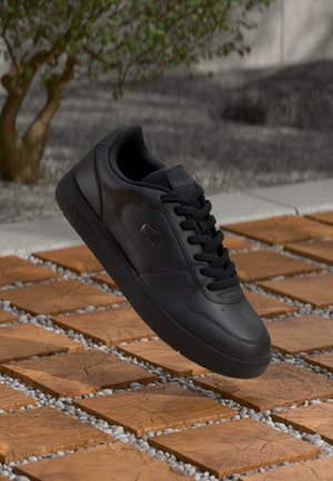Black leather sneaker with round toe, lace-up design, textured surface, and subtle branding. Placed above a patterned tile background.