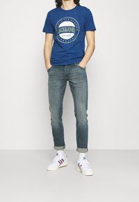 Man som bär en blå "Jack & Jones" T-shirt, uppvikta blekta blå jeans, vita strumpor och vita Adidas-sneakers med röda och blå ränder, stående med händerna i fickorna.