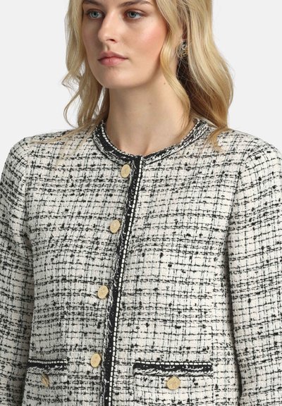 Bela in črna tvid jakna z okroglim ovratnikom, zlatimi gumbi in črnim okrasnim trakom. Ima dva našita žepa z kontrastnimi robovi.