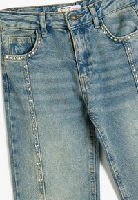 Jeans infantiles de denim azul con corte recto, adornados con brillantes remaches a lo largo de las costuras laterales y un ligero desvanecimiento en toda la tela.