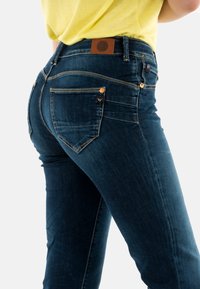 Vaqueros de mezclilla azul oscuro con un diseño ajustado, costuras en contraste, bolsillos en la parte posterior y un parche de cuero marrón en la cinturilla.