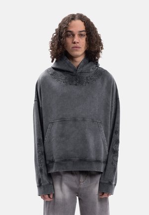 Ung person med krøllete hår som har på seg en falmet svart oversized hettegenser med subtil stammebroderi på krage og ermer, og grå jeans.