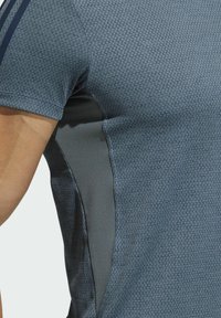 Primer plano de una persona que lleva una camisa de manga corta azul texturizada con un panel de malla gris oscuro en el lado debajo del brazo.