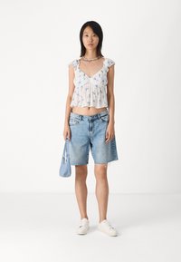 Bílý květinový crop top s volánovými rukávy, spárovaný se světle modrými denimovými šortkami a bílými teniskami. Malá modrá kabelka dotváří celkový vzhled.