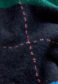 Wełniana tkanina w ciemnogranatowych i zielonych kolorach, z widocznym różowym ściegiem tworzącym wzór krzyża. Miękka w dotyku, o delikatnych włóknach.