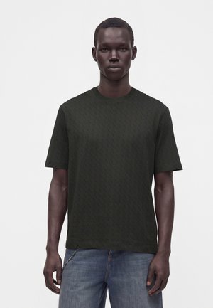 Jeune homme portant un t-shirt à manches courtes foncé à motifs et un jean bleu, debout devant un fond clair uni.