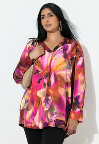 Kvinde med langt sort hår iført en livlig pink og orange blomstret bluse, sorte bukser, guldøreringe og holder en lille guldtaske.