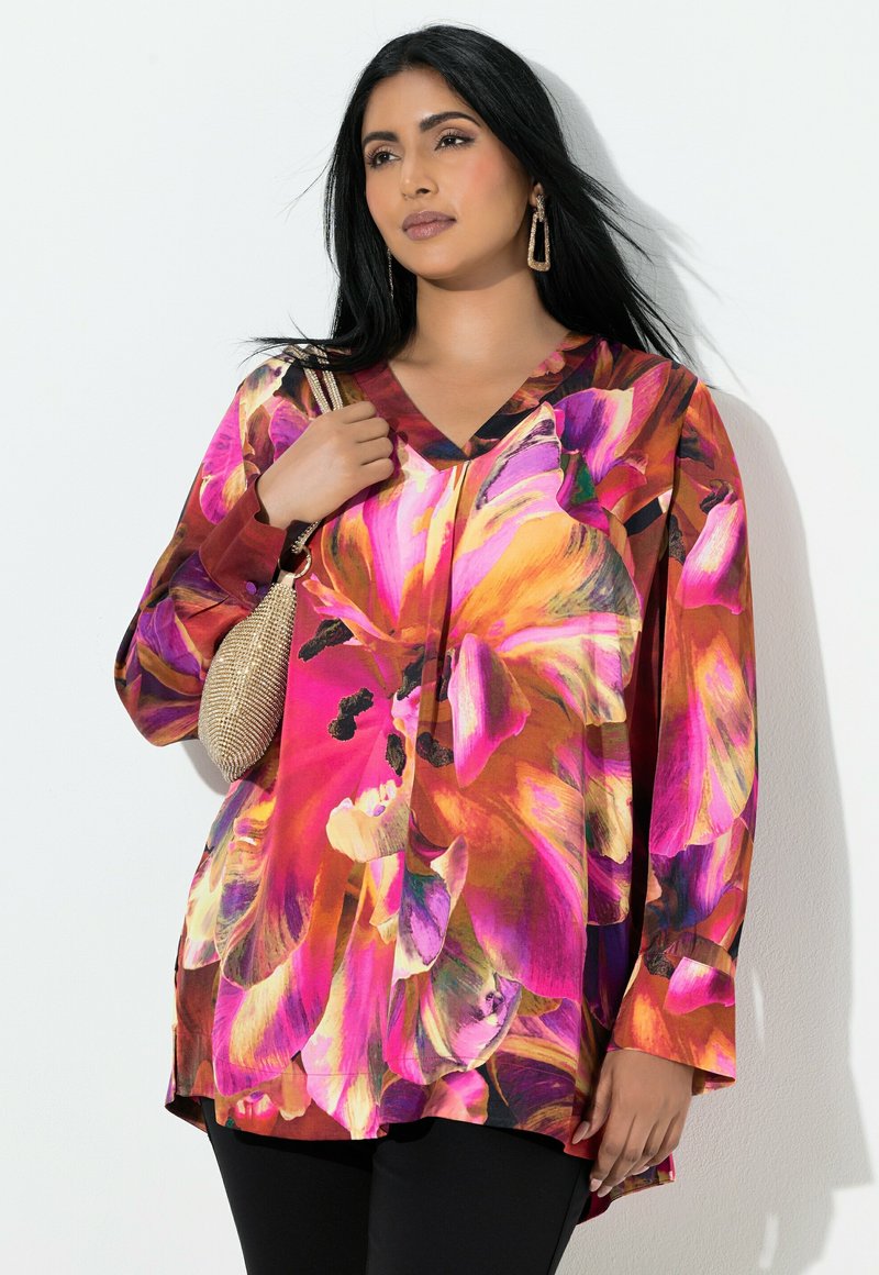 Kvinde med langt sort hår iført en livlig pink og orange blomstret bluse, sorte bukser, guldøreringe og holder en lille guldtaske.