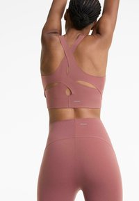 Woman with braided bun wearing mauve crisscross back sports bra and matching high-waisted leggings, arms raised above head.