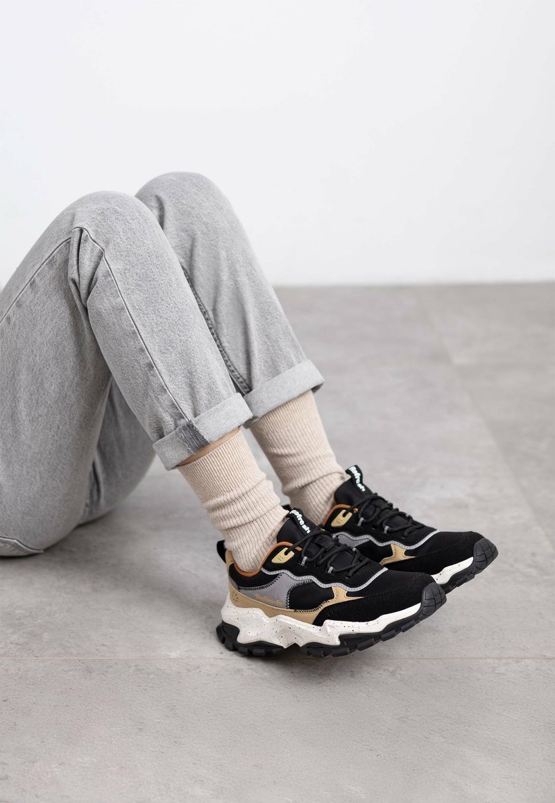 Zapatillas gruesas en negro y beige con tejido texturizado, que presentan un diseño ondulado y acentos grises en contraste. Colocadas sobre un fondo claro.