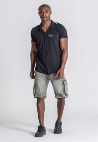 Camisa de botones de manga corta negra con botones dorados en los hombros, combinada con pantalones cortos cargo gris claro que cuentan con múltiples bolsillos. Zapatillas negras.