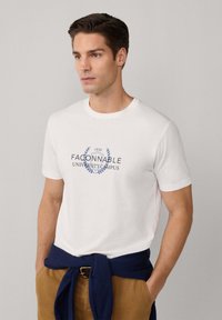 Mann trägt ein weißes T-Shirt mit dem Text "FACONNABLE UNIVERSITY CAMPUS", eine beige Hose und einen marineblauen Pullover, der um die Taille gebunden ist, und steht vor einem grauen Hintergrund.