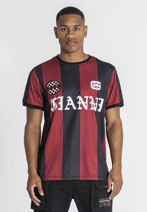 Jonge man die een rood-zwart verticaal gestreept shirt draagt met de tekst "Gianni", staand tegen een eenvoudige witte achtergrond.