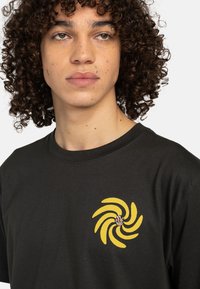 Schwarzes Baumwoll-T-Shirt mit einem gelben Spiralmotiv, das auf der linken Brust gedruckt ist. Klassischer Rundhalsausschnitt und kurze Ärmel.