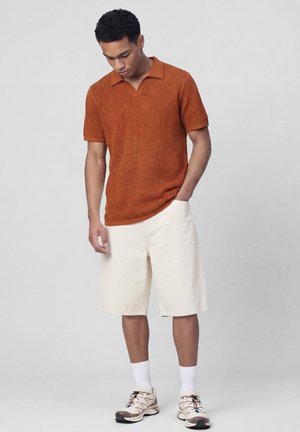 Homme en pull en maille à manches courtes de couleur rouille, short beige arrivant aux genoux, chaussettes blanches et baskets beiges, debout avec une main dans la poche.
