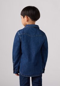 Camicia in denim blu scuro con colletto, maniche lunghe, polsi con bottoni e cuciture a contrasto. Presenta una spalla a yoke e un orlo curvo.