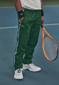 Pantaloni sportivi verdi con strisce laterali bianche e logo Lacoste, abbinati a scarpe da tennis bianche e grigie, e con in mano una racchetta da tennis arancione.