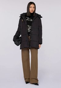 Svart vadderad jacka med huva, med kviltad design och knappar framtill. Kombinerad med lösa bruna byxor och en svart tröja.