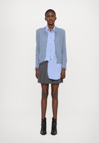Giacca blu con texture e bordi sfrangiati, camicia button-up azzurra chiara, gonna grigia plissettata e stivaletti neri alla caviglia con dettagli argentati.