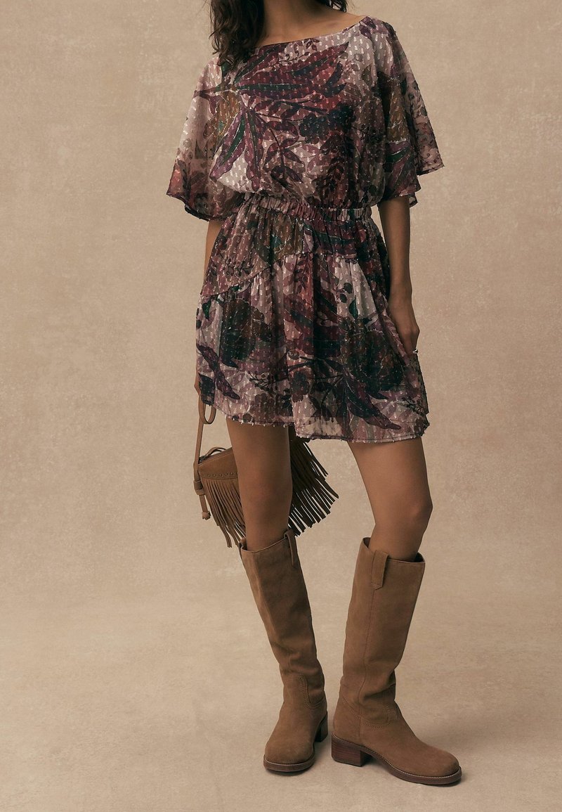 Femme portant une robe courte et transparente à motif floral avec une taille élastique, des bottes hautes en daim marron clair, et tenant un sac à main marron clair à franges.