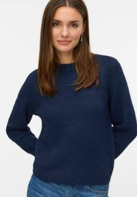 Tamno plavi teksturirani pulover s okruglim izrezom i dugim rukavima, uparen s plavim trapericama, prikazuje ležerni dizajn.