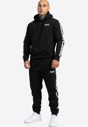 Zwarte katoenen hoodie met een voorzak en een capuchon met trekkoord, gecombineerd met bijpassende joggers met zijstreepdetails en logo's. Witte sneakers.