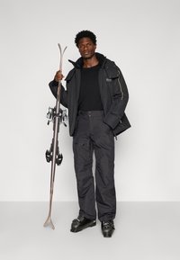 Casaco de esqui preto de inverno com capuz e bolsos com fecho, combinado com calças de esqui escuras e botas de esqui pretas, segurando esquis de madeira com ligaduras.