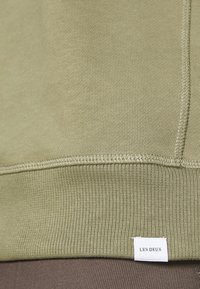 Orlo verde oliva chiaro con una finitura ribattuta e cuciture visibili, e una piccola etichetta tessuta bianca con la scritta "LES DEUX."