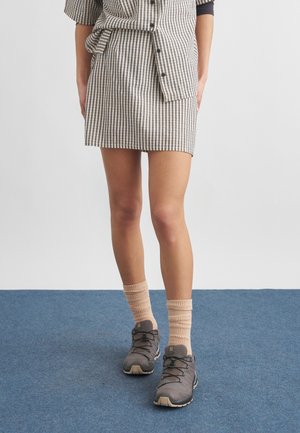 Persoon met beige geruite rok en overhemd, beige sokken en grijze wandelschoenen staand op een blauw tapijt.