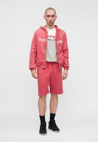 Homme debout portant un sweat à capuche zippé rouge, un short rouge assorti, un t-shirt blanc à motif graphique, des chaussettes noires et des baskets noires, sur un fond uni.